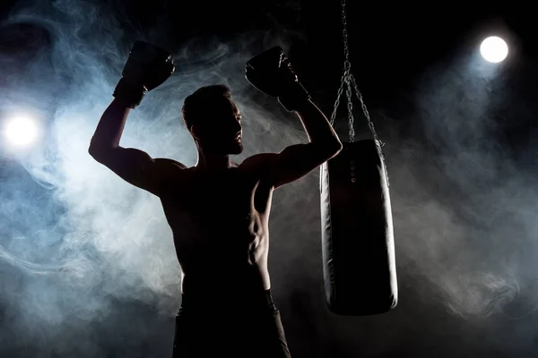 Silhouette of muscular athlete in boxing gloves with hands above head on black with smoke — Stock Photo