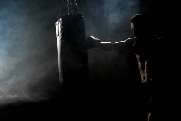 Silhouette of muscular athlete in boxing gloves kicking boxing bag on black with smoke — Stock Photo