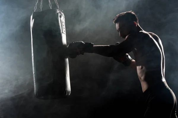 Silhouette of athlete in boxing gloves hitting punching bag on black with smoke — Stock Photo