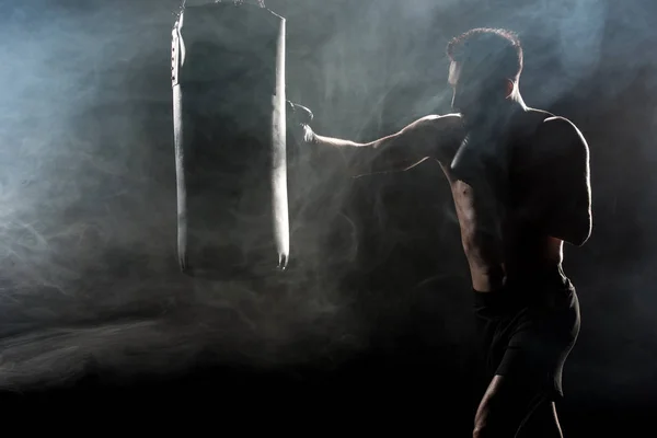 Silhouette of sportsman in boxing gloves hitting punching bag on black with smoke — Stock Photo