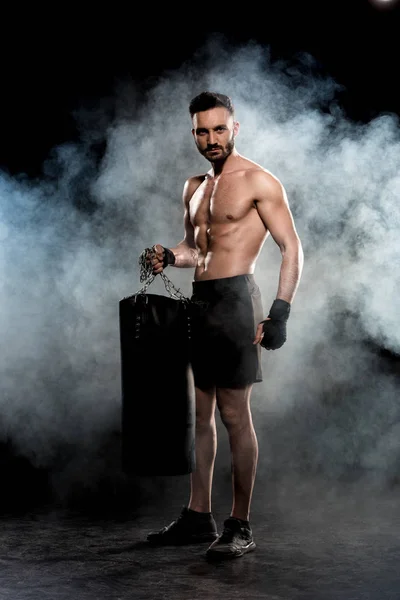 Handsome boxer holding punching bag on black with smoke — Stock Photo