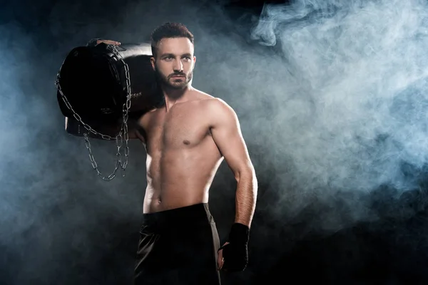 Boxer shortless segurando saco de perfuração no ombro no preto com fumaça — Fotografia de Stock