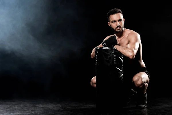 Hombre corto sentado y sosteniendo el saco de boxeo en negro con humo - foto de stock