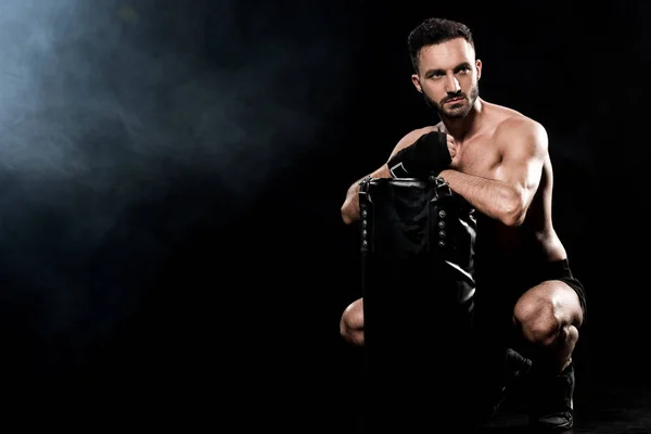 Shortless boxer sitting and holding punching bag on black with smoke — Stock Photo