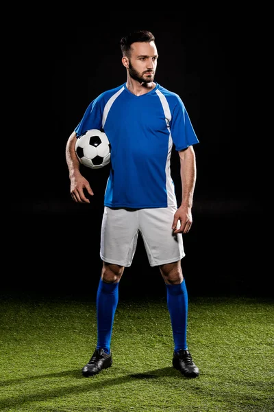 Apuesto jugador de fútbol de pie con pelota en hierba verde aislado en negro - foto de stock
