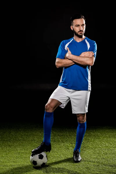 Jogador de futebol barbudo de pé com bola com braços cruzados isolados em preto — Fotografia de Stock