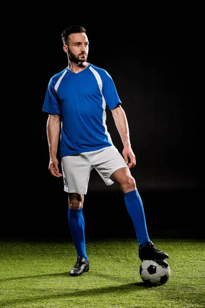 Barbuto giocatore di calcio in piedi con palla su erba isolato su nero — Stock Photo