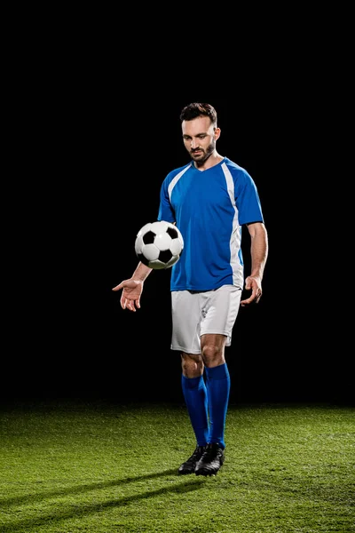 Bearded sportsman in uniform training with ball on grass isolated on black — Stock Photo