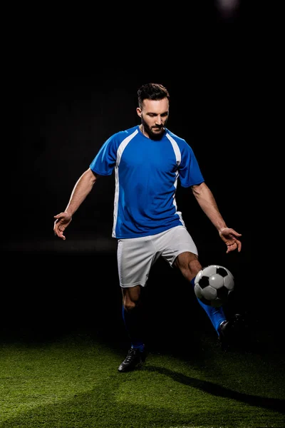 Jugador de fútbol barbudo en uniforme de entrenamiento con pelota sobre hierba aislado en negro - foto de stock