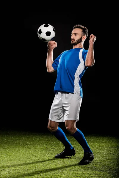 Futbolista atlético en uniforme saltando con pelota aislada en negro - foto de stock