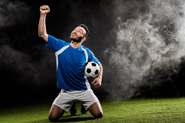 Jogador de futebol alegre comemorando a vitória e segurando bola no preto com fumaça — Fotografia de Stock