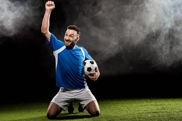 Happy football player celebrating victory and holding ball on black with smoke — Stock Photo