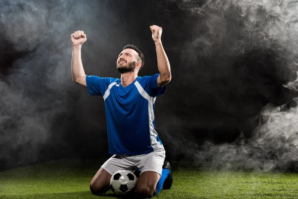 Jogador de futebol bonito comemorando a vitória enquanto sentado na grama em preto com fumaça — Fotografia de Stock