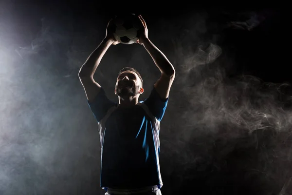 Silhouette of handsome football player holding ball above head on black with smoke — Stock Photo