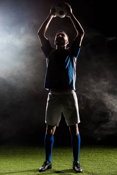 Силуэт футболиста, держащего мяч над головой на черном от дыма — стоковое фото