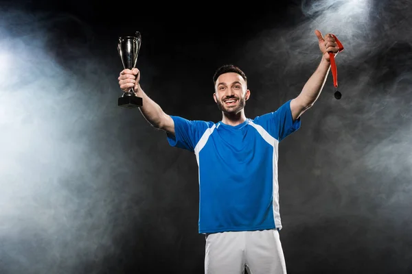 Guapo campeón sosteniendo trofeo y medallas en las manos en negro con humo - foto de stock