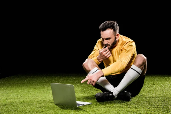 Arbitro guapo soplando en silbato y señalando con el dedo a la computadora portátil mientras está sentado en la hierba aislado en negro - foto de stock