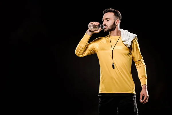 Handsome referee drinking water from bottle and standing with towel on shoulder isolated on black — Stock Photo