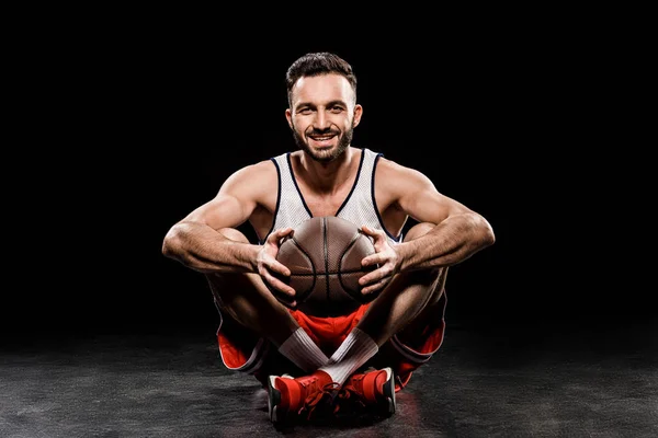 Fröhlicher Basketballspieler sitzt mit Ball auf schwarzem Hintergrund — Stockfoto