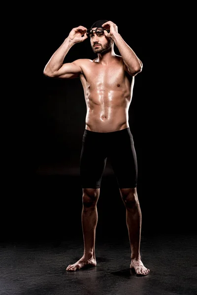 Muscular swimmer standing in swimming cap and touching swim goggles on black background — Stock Photo