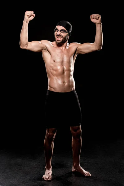 Nadador alegre de pie en gorra de natación y mostrando los músculos sobre fondo negro - foto de stock