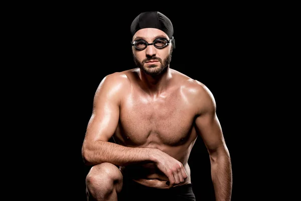 Muscular swimmer sitting in swimming cap and goggles isolated on black — Stock Photo