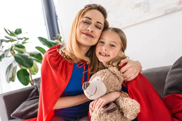 Hugging mother and kid in red cloaks holding teddy bear — Stock Photo