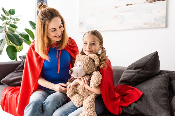 Красивая мать и ребенок в красных плащах держат плюшевого мишку и сидят на диване — стоковое фото