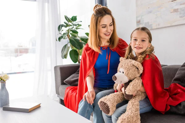 Beautiful mother and kid in red cloaks holding teddy bear and sitting on sofa at home — Stock Photo