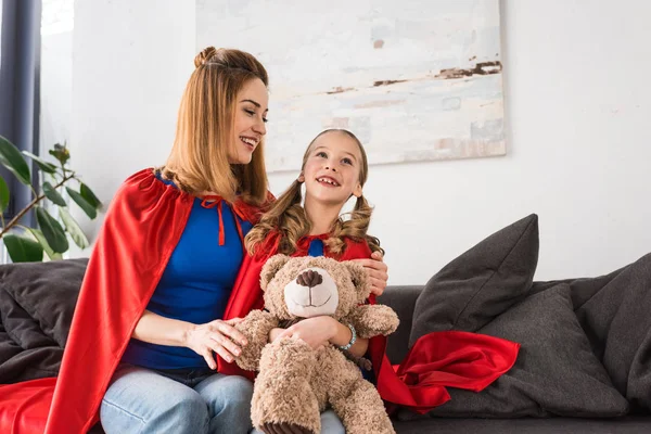 Madre e hija en capas rojas sonriendo y sosteniendo oso de peluche en casa - foto de stock
