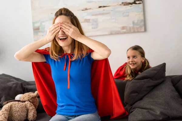 Sorridente madre e bambino carino in mantelli rossi giocare a nascondino a casa — Foto stock