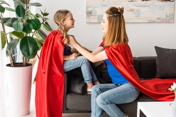 Hermosa madre atando rojo capa en hija en casa - foto de stock