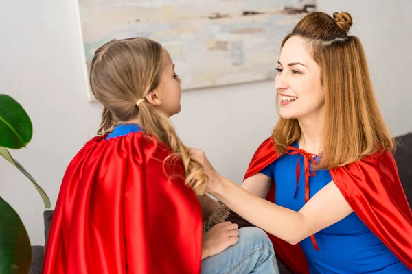 Cheerful mother tying red cloak on daughter at home — Stock Photo