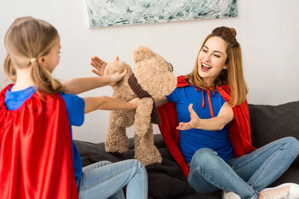 Beautiful mother and cute daughter in red cloaks playing with teddy bear at home — Stock Photo