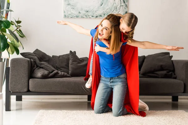 Hija y madre en capas rojas abrazándose y jugando en casa - foto de stock