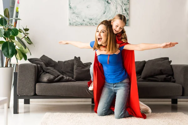 Filha e mãe em capas vermelhas sorrindo e brincando em casa — Fotografia de Stock