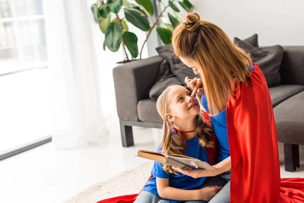 Daughter and mother in red cloaks sitting on floor and holding book — Stock Photo