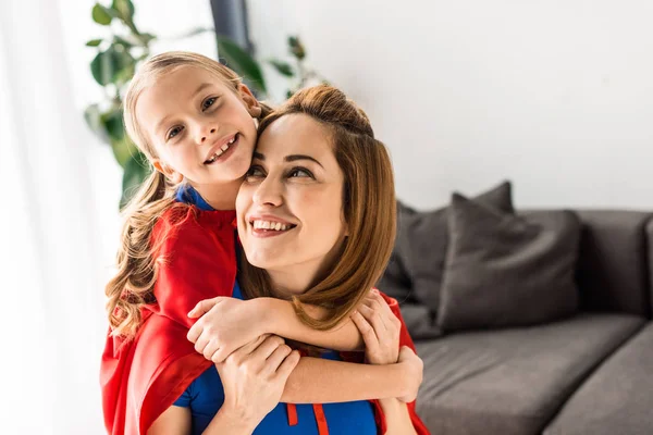 Linda hija y madre en capas rojas abrazando y sonriendo en casa - foto de stock