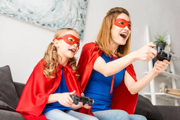 Alegre madre e hija en máscaras rojas y capas jugando videojuego en casa - foto de stock