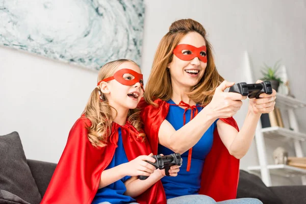 Sorridente madre e figlia in maschere rosse e mantelli che giocano al videogioco — Foto stock