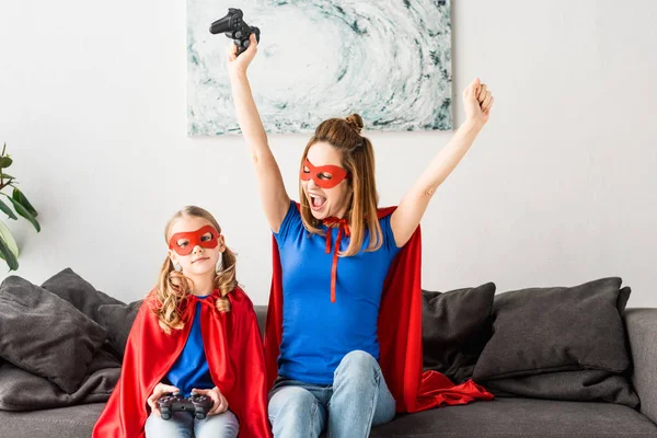 Atractiva madre e hija en máscaras rojas y capas jugando videojuego - foto de stock