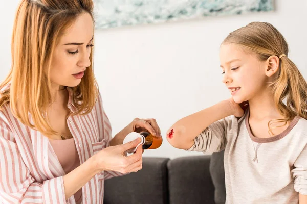 Attractive mother treating injured daughter with antiseptic at home — Stock Photo