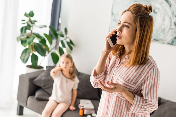 Upset mother talking on smartphone and daughter with wound sitting on sofa — Stock Photo