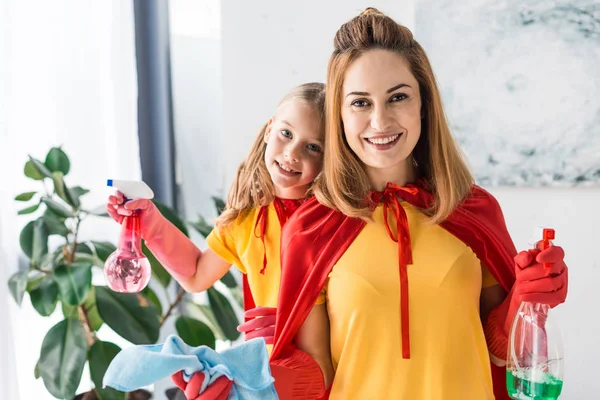 Mutter und Tochter in roten Umhängen und Gummihandschuhen mit Sprays reinigen und in die Kamera schauen — Stockfoto