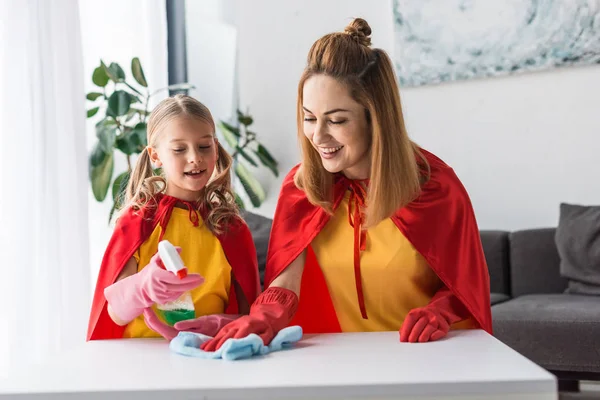 Mother and kid in red capes and rubber gloves dusting at home — Stock Photo