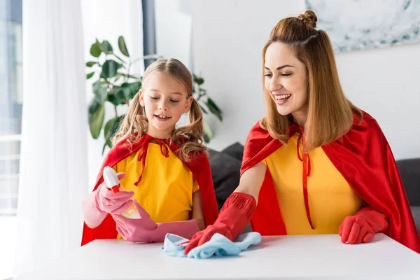 Мать и дочь в красных плащах и резиновых перчатках вытирают пыль дома — стоковое фото