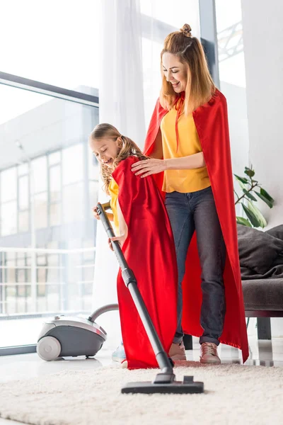 Mère et fille en capes rouges avec aspirateur à la maison — Photo de stock