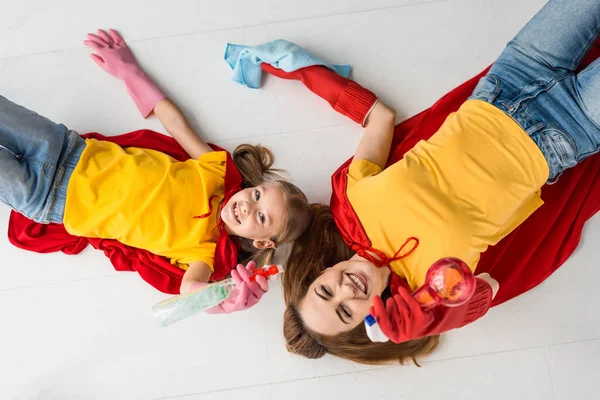 Vue du dessus de la mère et de l'enfant souriants en capes rouges et gants en caoutchouc avec pulvérisations à la maison — Photo de stock