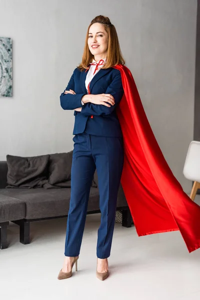 Femme d'affaires souriante avec les mains croisées en costume bleu et cape rouge regardant la caméra — Photo de stock