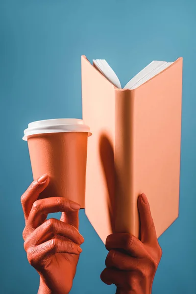 Vista recortada de las manos femeninas de color coral con taza de papel de coral y libro sobre fondo azul, concepto de color de 2019 - foto de stock